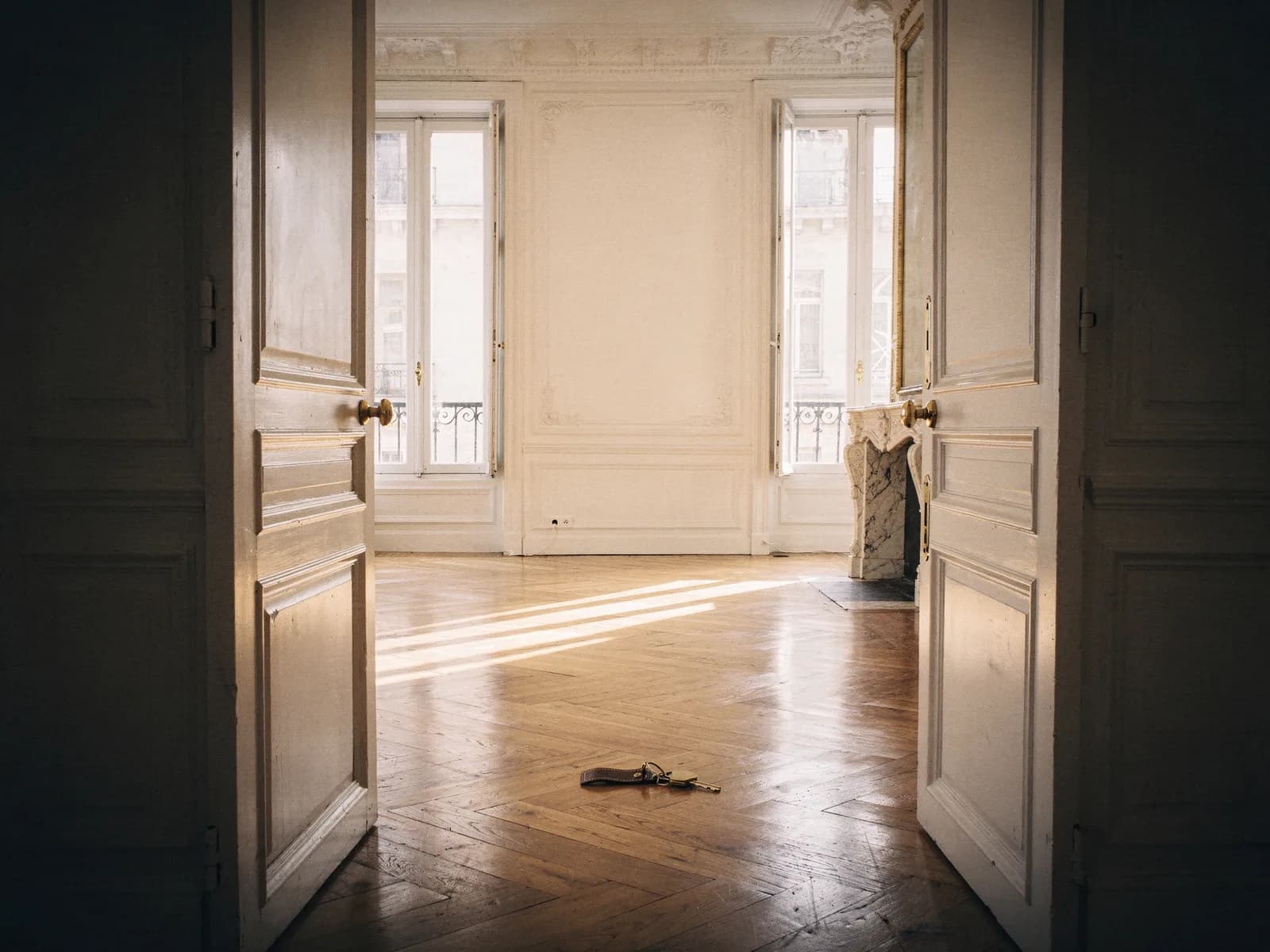 Illustration : par une double porte ouverte, une pièce haussmannienne vide et lumineuse, un trousseau de clés posé sur le parquet.