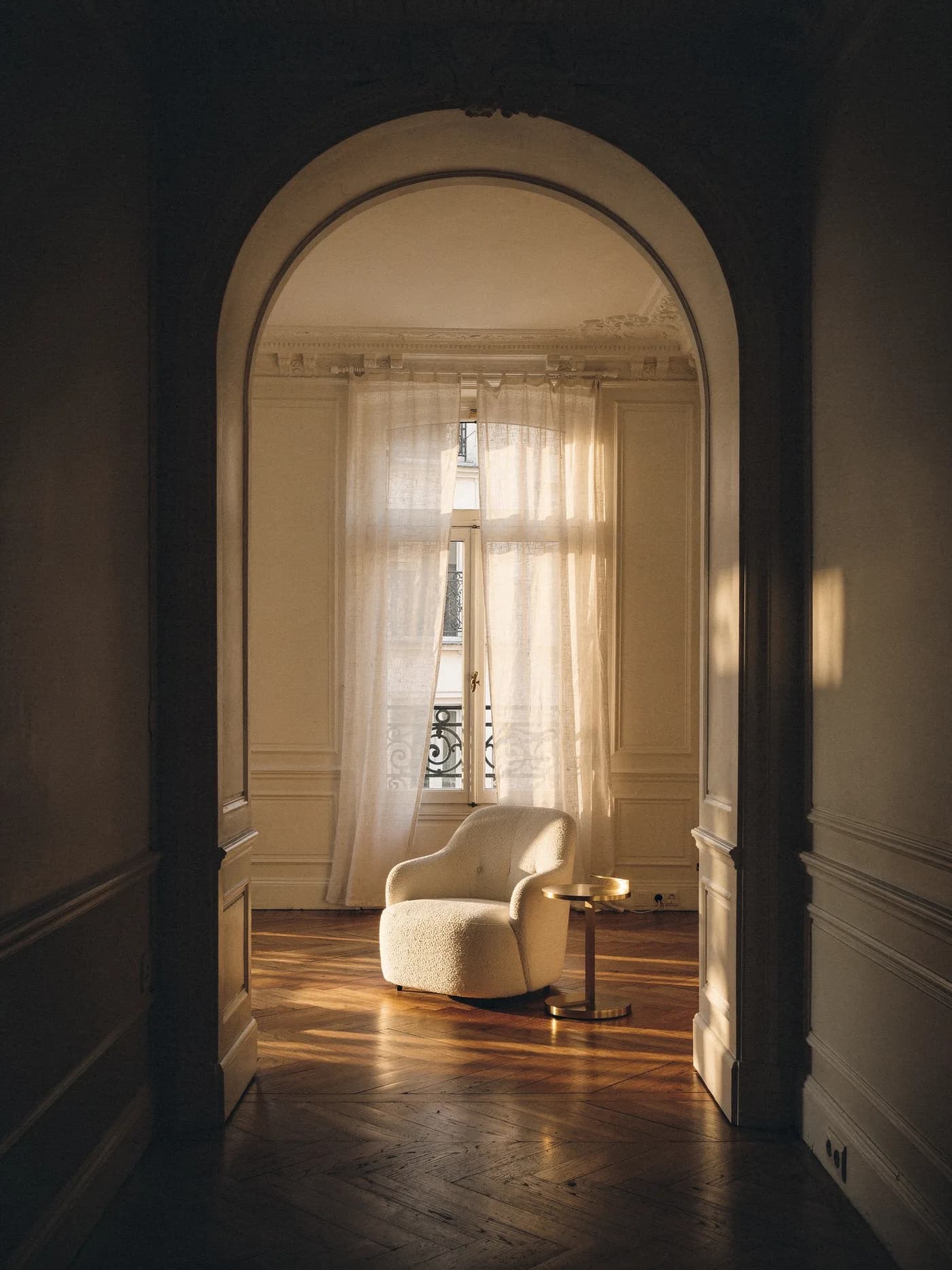 Illustration : depuis un couloir sombre, une arche ouvre sur un salon haussmannien baigné de lumière, un fauteuil clair près de la fenêtre.